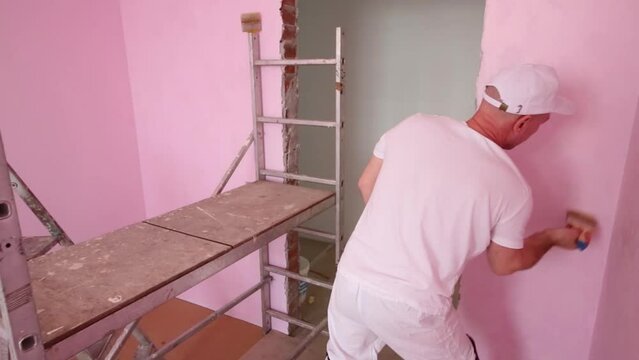 Worker in cap and white clothes paints wall in new room