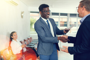 Happy middle-aged man holding a document shaking hands with a person in waiting hall. Visitor...
