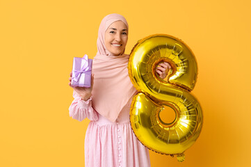 Mature Muslim woman with golden air balloon in shape of figure 8 and gift box on yellow background. International Women's Day