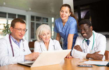 Three interested doctors sitting and a nurse standing at table searching information by means of notebook