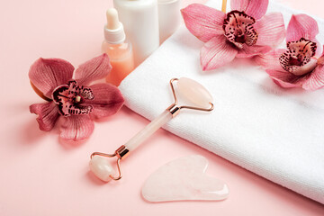 Facial cosmetic roller and gua sha scraper with pink orchid flowers on a white towel. Home spa concept