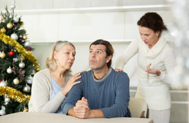 Woman is actively sorting out her relationship with man, and elderly woman is calming man while sitting at table in kitchen near christmas tree