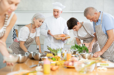 Positive middle-aged male cook conducting culinary classes for learners standing around table with piece of salmon in front of them
