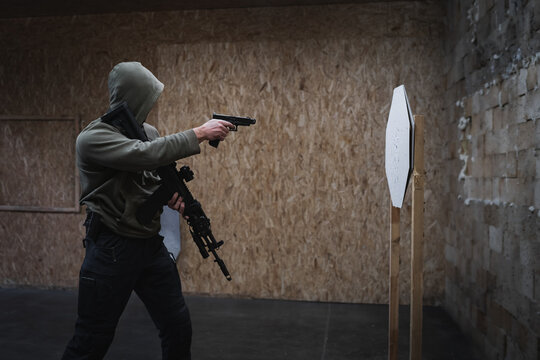 A man in a hooded sweater is engaged in tactical shooting with a pistol and rifle at a shooting range.