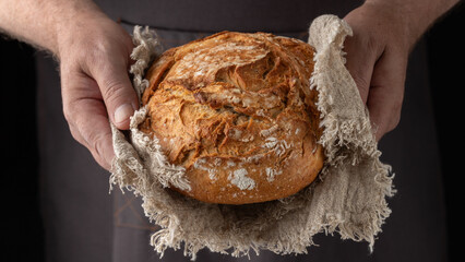 Man baker holding fresh spelt wheat loaf of bread on rustic linen towel on dark background. Healthy...