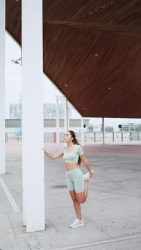 Vertical video of a fit young woman athlete warming up her legs before running outdoors in sportswear. Young sportswoman stretching during cardio training. Concept of sport, health and wellness.