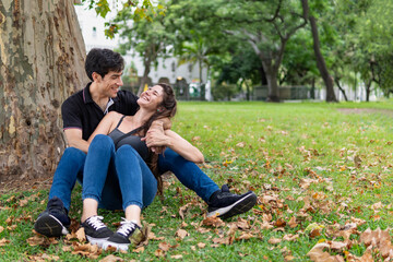 Couple sitting in a park leaning on a tree. Happy lovers laughing