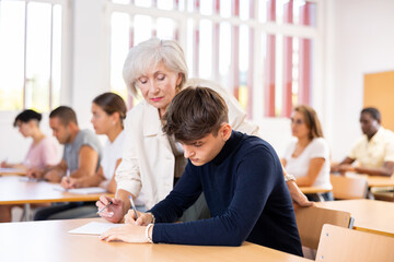 Experienced elderly female teacher helping to solve problem to young guy student sitting at desk,...