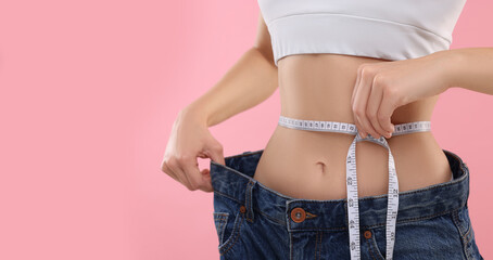 Slim woman wearing big jeans and measuring waist with tape on pink background, closeup. Weight loss