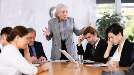 Irritated elderly female boss reprimanding upset subordinates sitting at table in office meeting room, expressing dissatisfaction with work