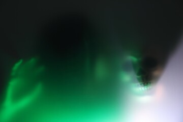 Silhouette of creepy ghost with skull behind glass against color background