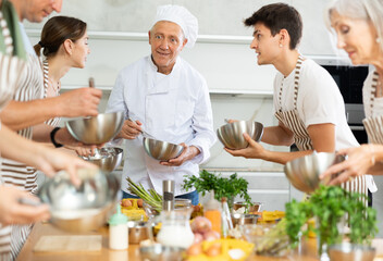Positive experienced senior chef giving culinary classes to group of interested men and women of different ages, teaching to cook deliciously..