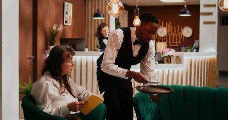 Bellhop serving coffee cup to woman client relaxing in lounge area, senior hotel guest enjoying...
