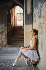 Ballerina in leotard and tutu on toes in sitting position against rustic wall.