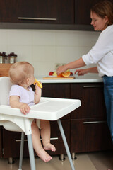 Adorable baby girl eating orange. Little daughter sitting in feeding chair and eating slice of orange at kitchen and playing with mom. Juicy fruits oranges are for a healthy diet. Happy childhood.