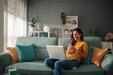Woman using laptop and typing on a phone while sitting on couch at home