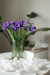 Concept of seasonal allergy to flower. Bouquet of purple iris flowers in a glass vase on table and many used napkins at home interior. Allergic rhinitis symptom of odor pollen flowering