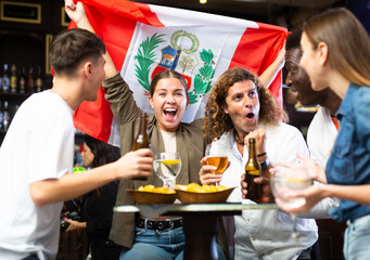 Cheerful diverse soccer supporters holding the flag of Peru and enjoing beer and potato chips, spending time together in the sport bar