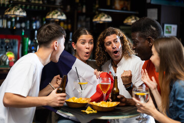 Group of cheerful young adult women and men having fun in sports bar, watching game on TV and rooting for favorite French team