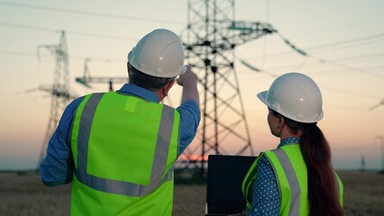 Woman electrician with laptop shows electric construction to man colleague. Electrical engineers discuss outcomes of day work at high voltage line tower. Employees share thoughts about lines condition
