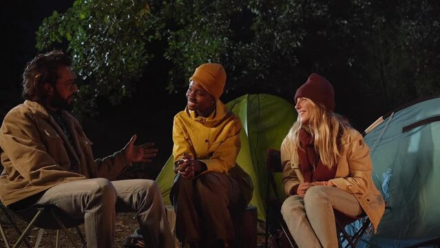 Three multicultural friends enjoying talking and laughing at winter night campsite getaway. Young cheerful group of persons together outdoors. Happy millennial people sitting at wilderness weekend.