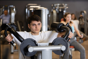 Fototapeta premium Focused young guy working out in modern gym, pumping up deltoid muscles of shoulders in lateral raise exercise machine. Active lifestyle concept and sports..