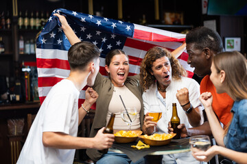 Cheerful diverse soccer supporters with flag of USA spending time together with pint of beer and...