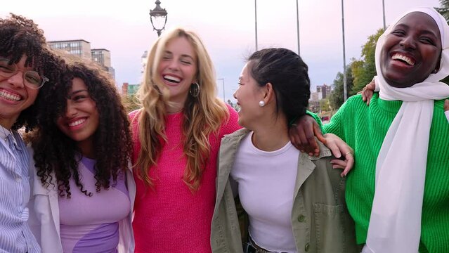 Seven happy young adult women from different cultures laughing together outdoors. Female friendship concept with diverse group of girls friends hugging each other having fun at city street
