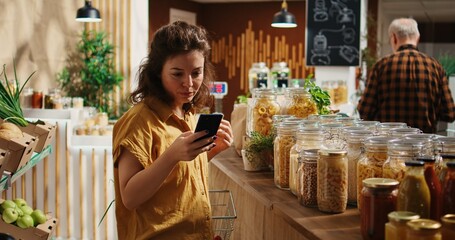 Woman using smartphone in zero waste supermarket to check what ingredients she needs for healthy recipe she plans to cook. Customer in local grocery shop uses phone while shopping for organic veggies