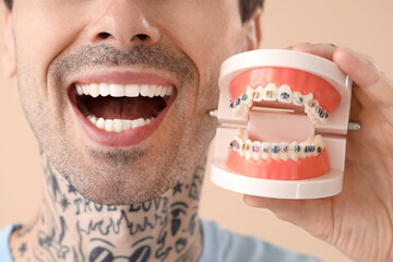 Young tattooed man holding jaw model with braces on beige background, closeup