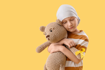 Cute little boy after chemotherapy with yellow ribbon and teddy bear on color background. Childhood cancer awareness concept - obrazy, fototapety, plakaty