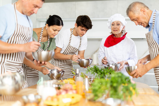Positive elderly woman professional chef conducting culinary courses, imparting cooking skills to diverse group of people of different ages..