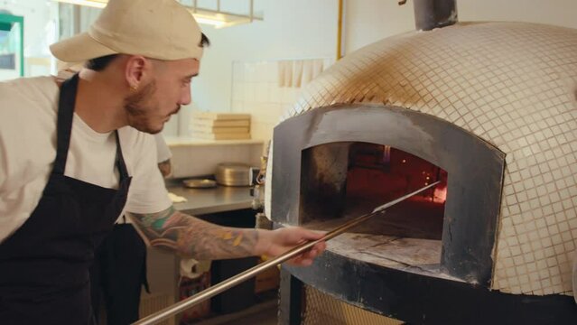 Chef using peel to turn coals and then starting fire in wood burning oven in the kitchen before opening pizzeria in the morning