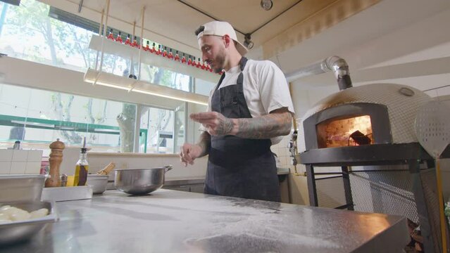 Professional chef in apron covering metal table with flour before working with dough in open kitchen during workday in pizzeria