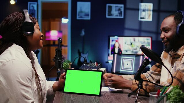 Woman and man participating in podcast, laughing while watching funny internet clips on green screen tablet in studio, discussing content. Podcasters reacting to amusing videos on chroma key device