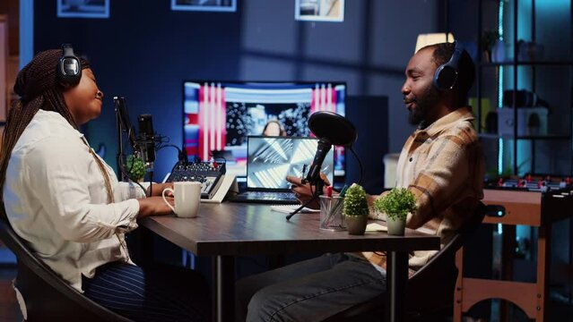 Online show host recording podcast with guest, using audio recording equipment in home studio living room. Happy man and woman laughing together during broadcasting session