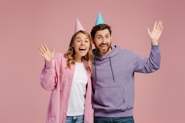 Couple of smiling attractive friends wearing birthday cone waving hand isolated on pink background....