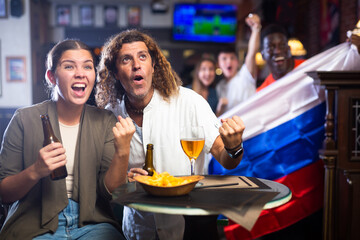 Excited diverse soccer supporters with flag of Russia watching tournament with pint of beer and...