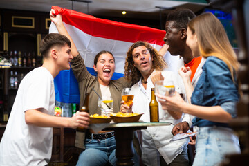 Group of excited young adults watching football championship in sports bar, supporting favorite team from Netherlands. People drinking beer with snacks at table and waving national Dutch flag..
