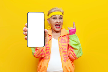 old funny grandmother in youth disco costume shows blank smartphone screen and rock gesture on...