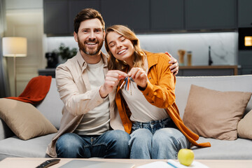 Happy lovely couple holding keys from new home looking at camera sitting on comfortable sofa. Smiling middle aged man and woman buying apartments - obrazy, fototapety, plakaty