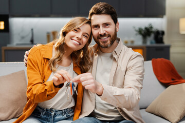 Portrait of happy young family holding keys from new home. Attractive man and woman hugging buying new property sitting together on comfortable sofa. Moving concept