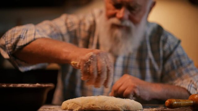 Close-up. An elderly man with a beard in a shirt sprinkles flour with his hand on an oblong piece of dough lying on a wooden table. A man cooks while sitting at a table in the kitchen in a modern home