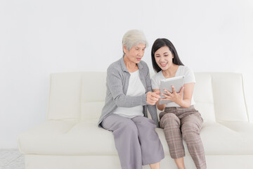 Obraz premium young Asian female and old female search information with tablet computer, they feeling happy and smile, they sitting on sofa, mother's day and happiness family time, technology and social network