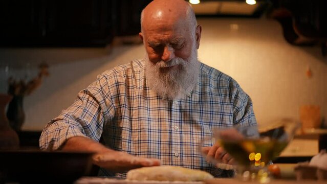 Close-up. An elderly bald man with a beard in a shirt pours vegetable oil, from a glass container with a handle, onto an oblong piece of dough and rubs it with his hand. The dough lies on a wooden