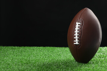 American football ball on green grass against black background, space for text