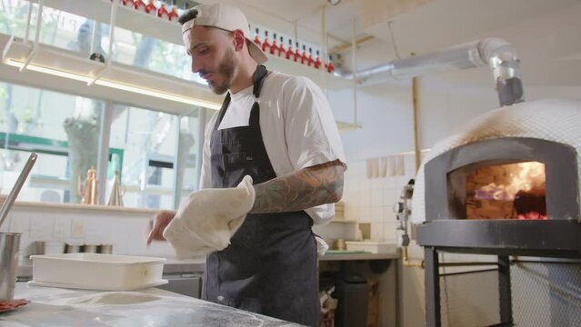Chef tossing and stretching pizza dough when his coworker keeping fire in wood burning oven during workday in open kitchen at pizzeria