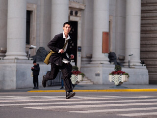 Portrait of handsome Chinese young man with black short hair wearing black blazer running across street with modern city building background in sunny winter day, male fashion, cool Asian young man.