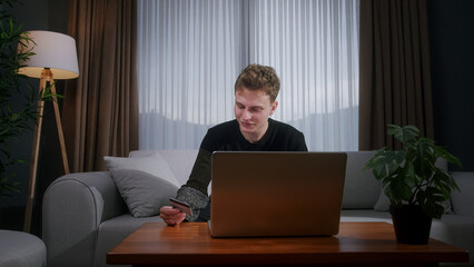 Smiling young man sitting enter credit card number on laptop for makes secure easy distant electronic payment while sitting on sofa at home. Pay for goods services on internet, transactions	
