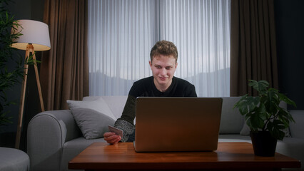 Young blonde man sitting enter credit card number on laptop for makes secure easy distant electronic payment while sitting on sofa at home. Pay for goods services on internet, transactions	
